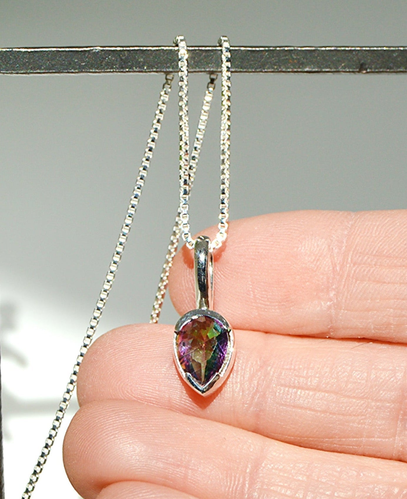 Teardrop-shaped gemstone pendant on a chain held by a hand against a neutral background